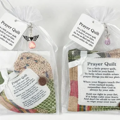 Two prayer quilts in clear bags, each with an attached charm and informational tag.
